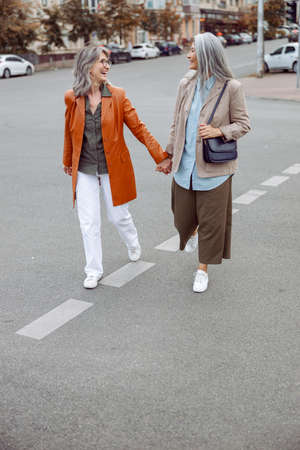 Happy senior women walk joining hands along modern city street on autumn dayの写真素材