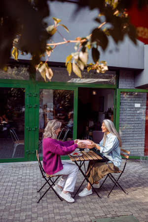 Positive senior women companions hold gift box sitting table on cafe terraceの写真素材