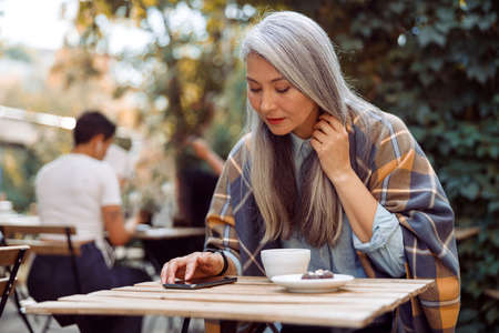 Long haired mature Asian lady reads message on mobile phone sitting on outdoors cafe terraceの写真素材
