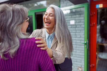 Emotional mature Asian lady laughs meeting grey haired best friend on modern city streetの写真素材