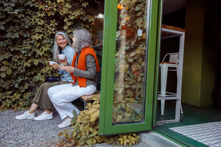 Cheerful senior ladies friends with cups of drinks sit on bench in cafe yardの写真素材