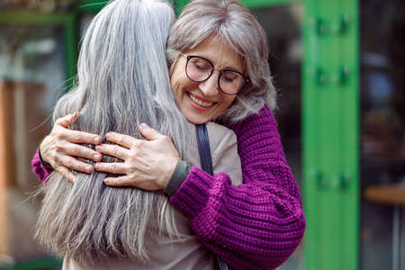 Smiling mature woman embraces female friend meeting on modern city streetの写真素材