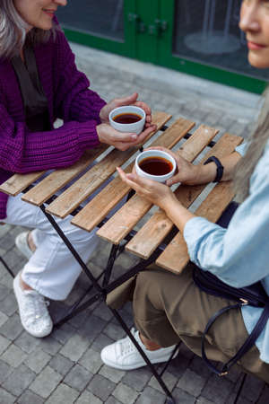 Senior ladies old friends hold cups of coffee sitting at small table on outdoors cafe terrace