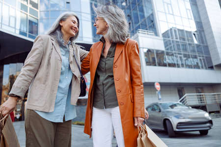 Smiling grey haired Asian woman hugs mature lady holding shopping bags on city streetの写真素材