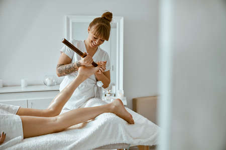 Conident female specialist is doing leg massage with bamboo stick to beautiful caucasean woman clientの写真素材