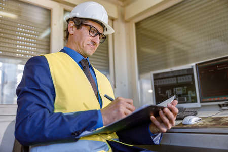 Male engineer in work vest writing documents at factory