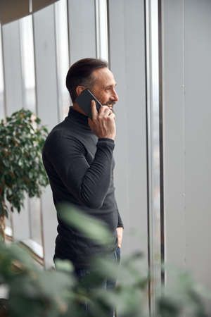 Happy smart adult caucasian businessman is standing near big windows in office space and holding...