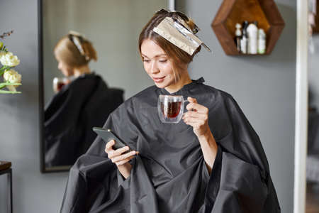 Happy blonde caucasian woman is using smartphone while sitting in chair and waiting in hairdressers...