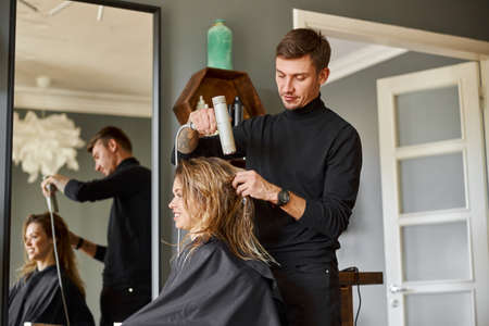 Happy beautiful light hair caucasian woman at beauty salon. Male master is drying her hairの写真素材