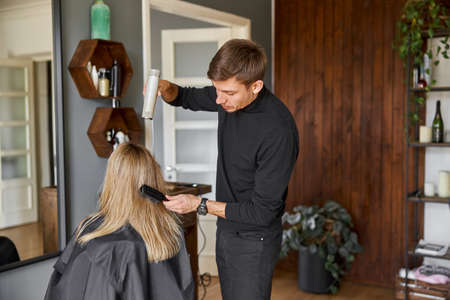 Happy beautiful light hair caucasian woman at beauty salon. Male master is drying her hairの写真素材