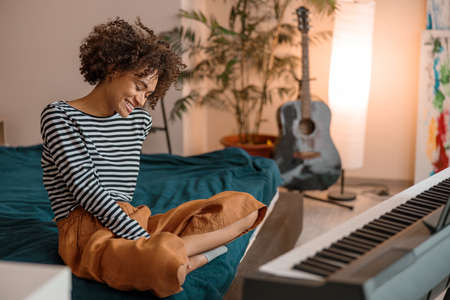 Joyful African woman sitting near synthesizer at homeの写真素材