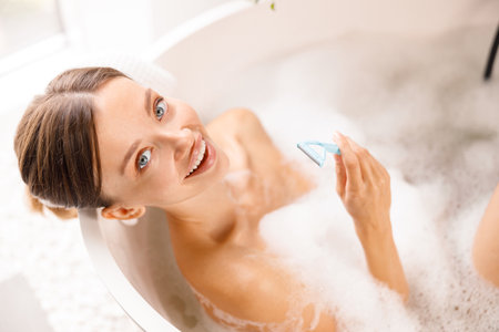Happy young woman smiling at camera, holding disposable shaving razor for shaving her legs in the bath with foamの写真素材