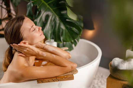 Peaceful young woman leaning on bathtub side andresting in the bathroom decorated with tropical plantの写真素材