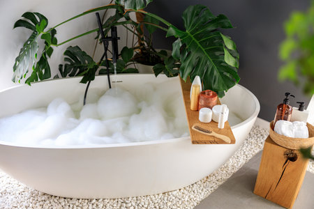 Modern bathroom interior with white bubble filled bathtub and monstera tropical plant. Eco body care products on wooden home decor elementsの写真素材