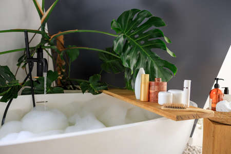 Wooden shelf for beauty and body care products on wooden shelf over modern white bubble filled bathtub. Cozy stylish bathroom decorated with green tropical plant Monsteraの写真素材
