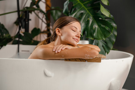Dreamy young woman leaning on bathtub side and resting in the bathroom decorated with tropical plantの写真素材