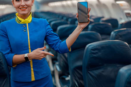 Smiling stewardess with mobile phone standing in airplane cabinの写真素材