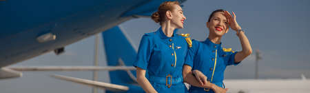 Two cheerful air stewardesses in bright blue uniform walking outdoors in front of passenger aircraft...