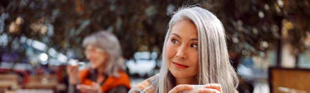Senior cafe guests, focus on dreamful long haired mature lady holding cup of drink at table