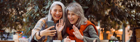 Positive mature women friends wave hands at videochat via smartphone in street cafe