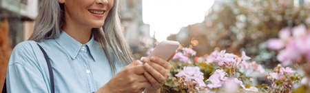 Smiling grey haired Asian woman uses cellphone standing on outdoors terraceの写真素材