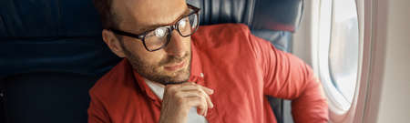 Handsome caucasian man in casual wear and glasses looking thoughtful, sitting on the plane near the windowの写真素材
