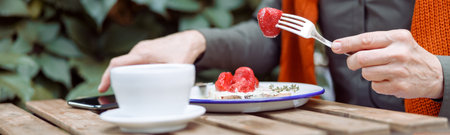 Mature woman eats toast with cream and strawberries taking mobile phone at table on outdoors