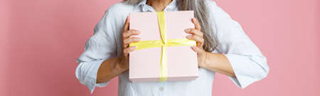 Emotional Asian woman with loose grey hair and party hat holds present on pink backgroundの写真素材