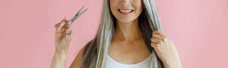 Cheerful Asian lady holds lock of silver hair and scissors on pink backgroundの写真素材