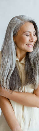 Shying middle aged Asian lady with long silver hair poses on light grey backgroundの写真素材