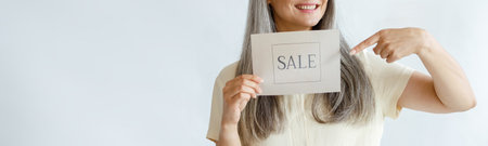 Joyful middle aged Asian woman with grey hair points onto Sale sign on light backgroundの写真素材