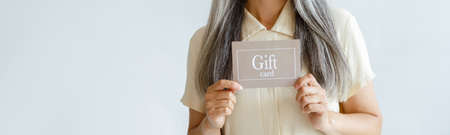 Smiling grey haired Asian woman holds gift card standing on light backgroundの写真素材