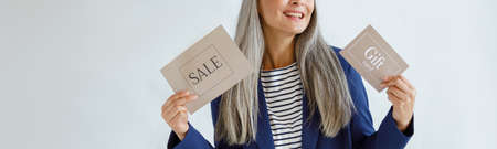 Cheerful middle aged Asian woman holds gift card and Sale sign standing in studioの写真素材