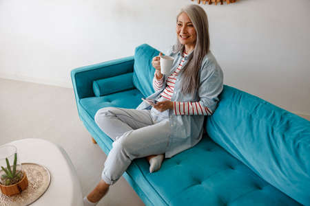 Joyful woman drinking coffee and using smartphone at homeの写真素材