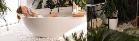 Relaxed young woman bathing in modern bathroom interior decorated with tropical plants