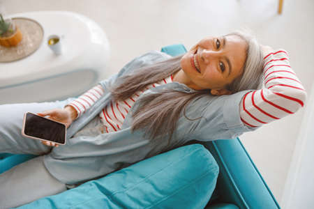 Cheerful woman with smartphone lying on couch at homeの写真素材