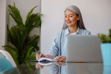 Cheerful woman writing in notebook and using laptop at homeの写真素材