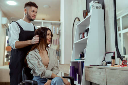 Beautiful woman with long hair looking pleased while hairstylist doing her hair at beauty salonの写真素材