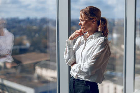 Caucasian businesswoman standing in office and looking out of window. Mature woman looking awayの写真素材