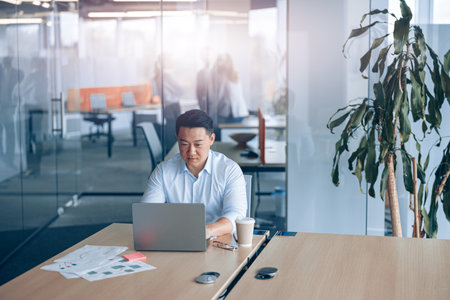 Attentive asian businessman working on laptop at workplace in modern office