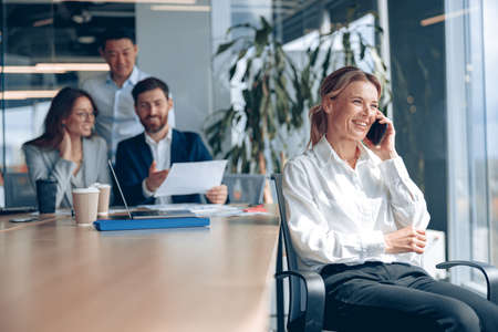 smiling mature lady is sitting on a chair and talking on phone during meeting in modern officeの写真素材
