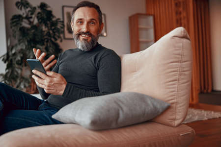 Smiling bearded man with mobile phone while sitting in cosy armchair