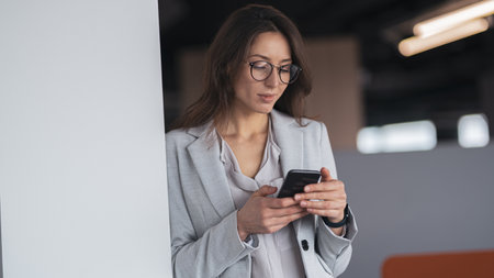 Focused Businesswoman wearing glasses with mobile phone in hand standing in modern office