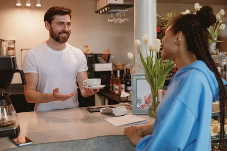 Making and handing coffee for young woman at counter in cafeteria. Happy waiter at bar.の写真素材