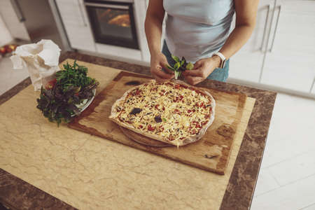A woman adds basil to the pizza, cooking at modern kitchenの写真素材