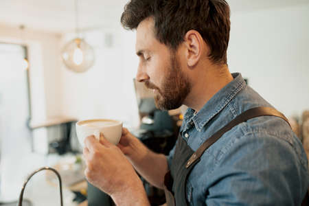 Handsome barista with stylish beard smelling coffee in cafeteria