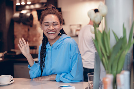 Pretty mixed-race young female bartender with piercing smiling at counter and waving hand.の写真素材