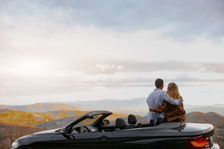 Happy couple man and woman enjoying beautiful view traveling in cabriolet looking at mountainsの写真素材