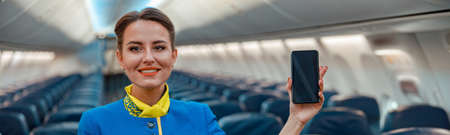 Joyful stewardess with smartphone standing in airplane cabinの写真素材