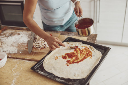 A woman spreads tomato sauce on a pizza laid out on the oven for bakingの写真素材